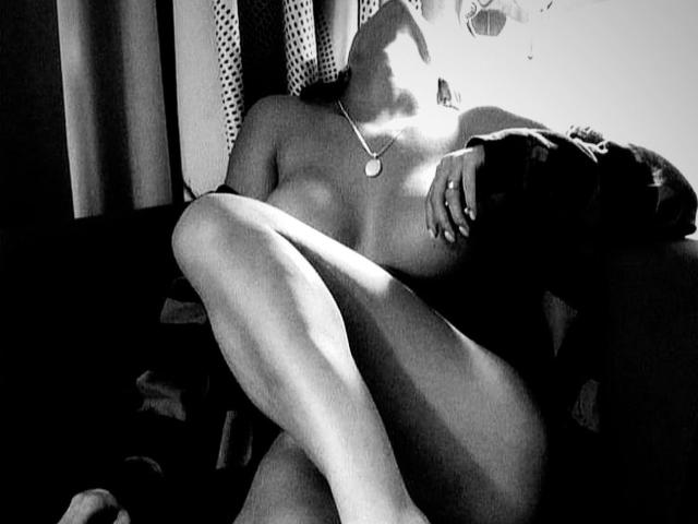 Black-and-white photo of Lisabet, naked, with legs crossed, small tits, a necklace, and dark hair, sitting in shadowy, dimly lit room, hand on chest, looking up.