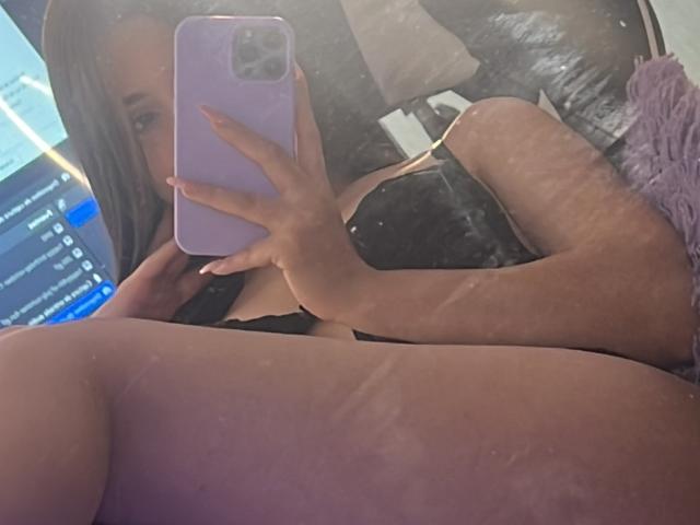 Nikole Foster, con cabello castaño largo, un sostén negro y un teléfono morado, está tomando una selfie frente al espejo, mostrando sus muslos desnudos y el reflejo de su rostro. Su mano cubre sus pechos..