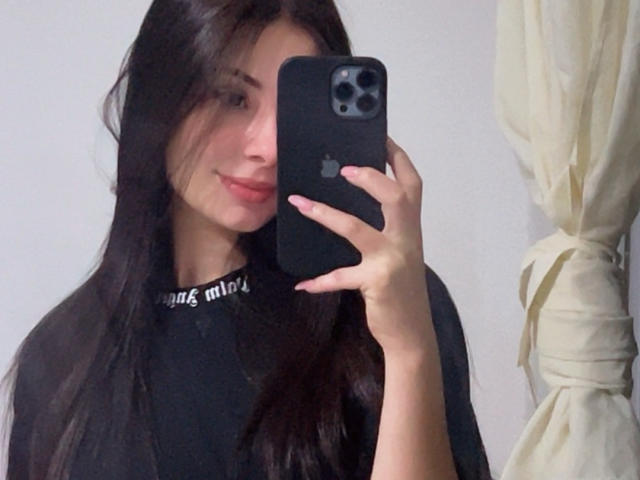 Nikole Foster, avec de longs cheveux foncés, un haut noir, tenant un iPhone noir, prenant un selfie, souriant légèrement, avec des ongles roses, un mur blanc, un rideau beige sur la droite..