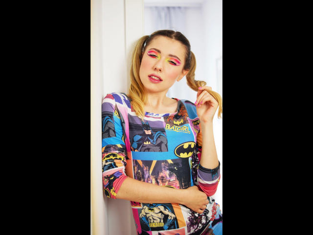 FlexiHeidi-hot with blonde pigtails, wearing a colorful Batman-themed shirt, has pink eyeshadow and red lipstick, standing against a white wall, holding her hair.