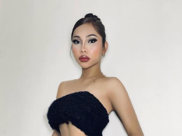 PerfectTransCum with dark brown hair in a bun, wearing a black fuzzy strapless top, has full lips, dark eyeliner, and small hoop earrings, standing against a plain white background.