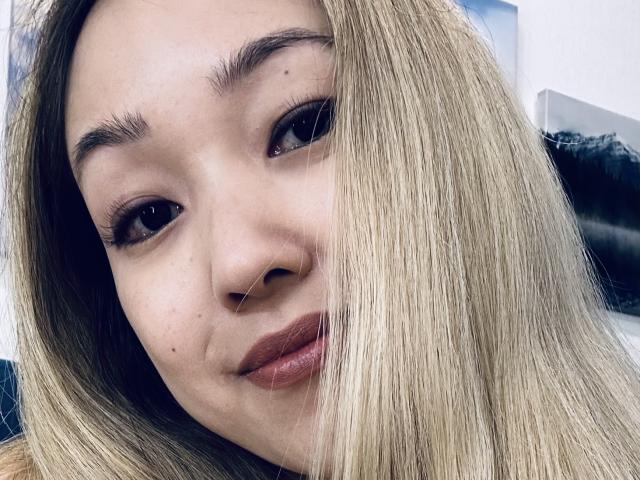 RenyLime's face, blonde hair covering one eye, brown eyes, thick eyebrows, subtle smile, light brown lipstick, small moles, slightly parted lips, soft skin, close-up selfie.