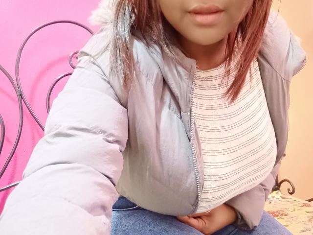 Brenadahy, avec des cheveux brun-rosés, portant une veste matelassée gris clair et une chemise blanche rayée, montrant de larges seins, assise avec une main sur le ventre, un mur rose en arrière-plan..