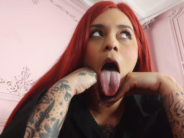 Chili with red hair and tattoos, sticking her tongue out, her finger touching her cheek, eyes wide and mouth open.