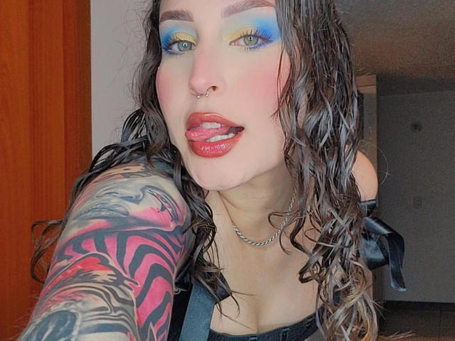 RoseCreamX with wet brown hair, blue-yellow eyeshadow, nose ring, red lipstick, tongue out, black strap dress, and a colorful tiger tattoo on her left arm.