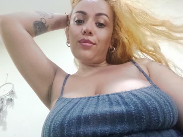 Joanallen with blonde hair, wearing a blue ribbed top, showing big tits, tattoo on arm, earrings, and a slight smile, looking down at the camera.