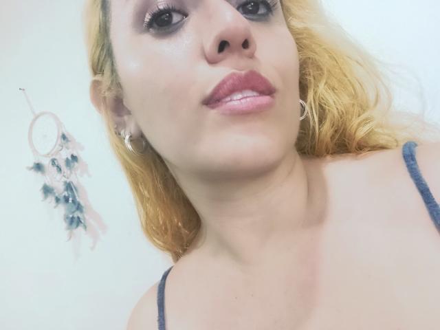 Joanallen with blonde hair, pink lipstick, and hoop earrings, wearing a blue top, has shiny skin and full pink lips, showing a slight pout, with visible cleavage.