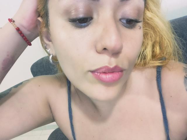 Joanallen a la peau pâle, du rouge à lèvres rose et des cheveux blonds. Elle porte un haut noir à bretelles fines et regarde vers le bas avec les yeux à moitié fermés. Elle a un bracelet rouge à son poignet gauche..