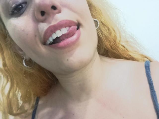 Joanallen's face, showing pale skin, blonde wavy hair, hoop earrings, and pink lips with tongue out, wearing a blue spaghetti strap top, showing her small tits.