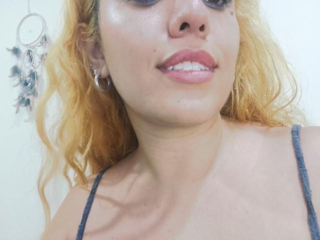 Joanallen has light skin, wavy blonde hair, and is smiling with pink lipstick. She's wearing a grey top and hoop earrings, showing her shiny, slightly sweaty face.
