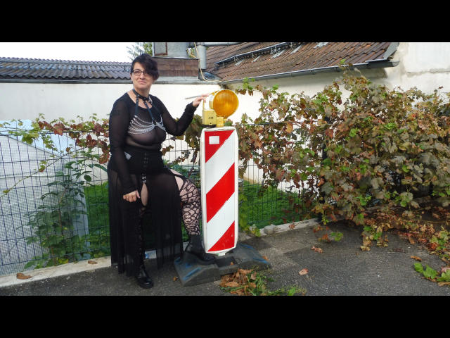 EinfachDani-hot with tits, wearing black sheer top, fishnet pants, and boots, sitting next to a red and white striped road sign, holding a yellow reflector.