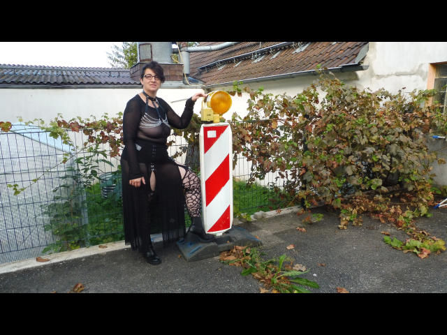 EinfachDani-hot with big tits, black sheer top, black fringed shawl, black shorts, holding yellow traffic light, standing by red-white striped barrier on pavement.
