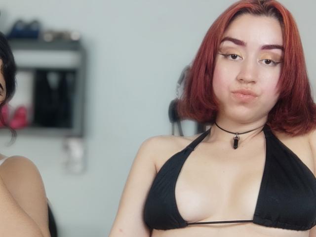 BelindaAndEmily with red hair, black bikini top showing small tits, black choker, and subtle makeup, looking at the camera with a slight pout. Partial view of another person.