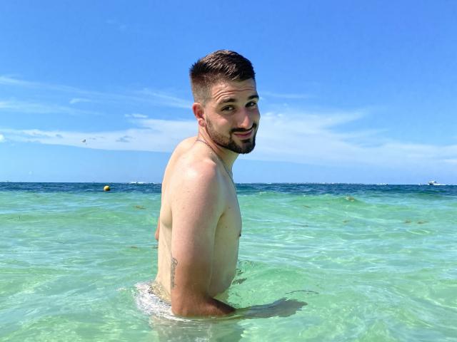 NaughtyGuyX, shirtless, standing in clear blue-green water, looking back with a slight smile, short brown hair, trimmed beard, tattoo on right arm, bright sunny day, clear sky.