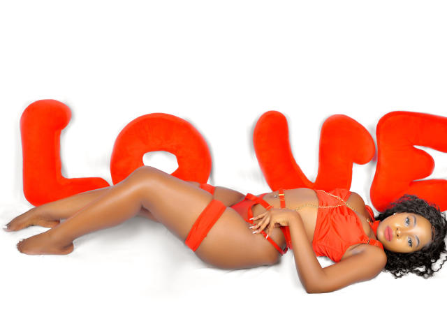 PrettyRebecca lies on her back, wearing red lingerie with a garter. She has dark skin, curly black hair, and a slim, sexy body with small tits and a cute ass.