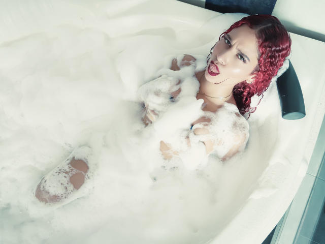 ValeryScot with red curly hair, naked, in a white bathtub filled with thick, white foam, smiling with tongue out, visible boobs and ass, partially covered by foam.