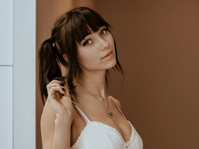 LuciaRare-hot with puffy brown tits, wearing white lace bra, brown pigtails, holding hair, green eyes, subtle smile, necklace with green pendant, soft brown background.