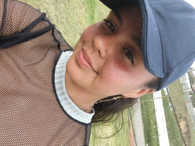 ArielleWilde with brown skin, wearing a mesh top showing her tits, blue cap, gold earrings, and a subtle smile, standing outdoors with grass in the background.