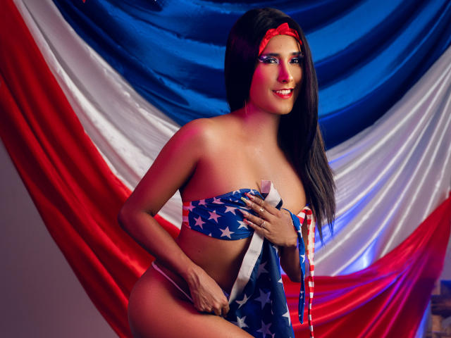 FallonRogers mit langem, schwarzem Haar, die ein rotes Stirnband, ein blaues Bikini-Oberteil mit Sternenmuster und eine amerikanische Flagge im Hintergrund trägt, hält die Flagge, zeigt ihre Brüste, lächelt, mit roter und blauer Beleuchtung..