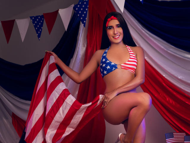 FallonRogers mit Bikini mit amerikanischer Flagge, hält eine weitere Flagge, rotes Stirnband, lächelnd, Knie angezogen, Hintergrund in Rot, Weiß und Blau, Flaggen in Rot, Weiß und Blau, mittelgroße Brüste, gebräunte Haut, langes, dunkles Haar..