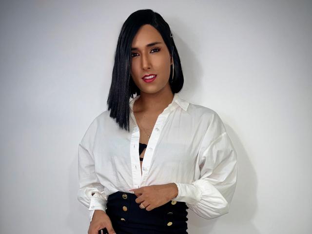 FallonRogers con caschetto nero, camicia bianca con bottoni, reggiseno nero e pantaloni neri a vita alta, che mostra leggermente il seno, sorridente, che tiene i pantaloni, su sfondo bianco..