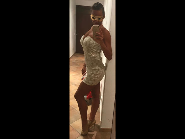 LorenaBella, atractiva con piel morena, vestida con un vestido ajustado, brillante y sin tirantes, gafas amarillas y tacones, posando en un selfie frente a un espejo, mostrando su trasero y sus pequeños senos firmes..