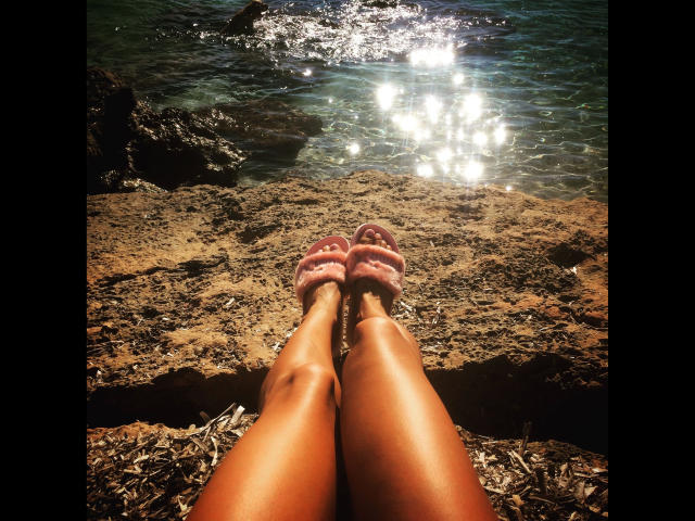 LorenaBella: gambe abbronzate e dalla pelle luminosa, piedi in sandali rossi, appoggiate su una spiaggia rocciosa, l'acqua che brilla alla luce del sole, le gambe rivolte verso l'oceano scintillante..