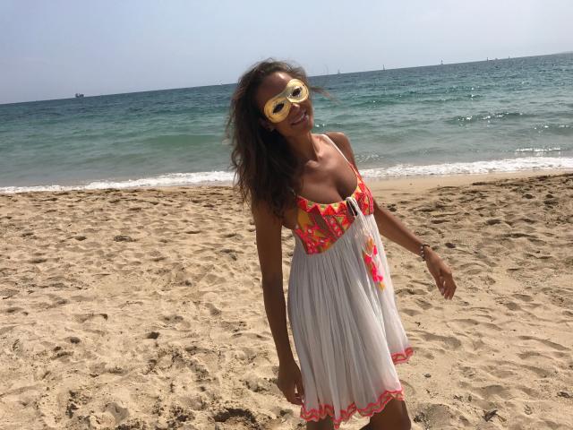 LorenaBella-hot in a white, floral dress with pink accents, yellow sunglasses, and wavy brown hair, standing on a sandy beach, her small tits and cute ass visible.