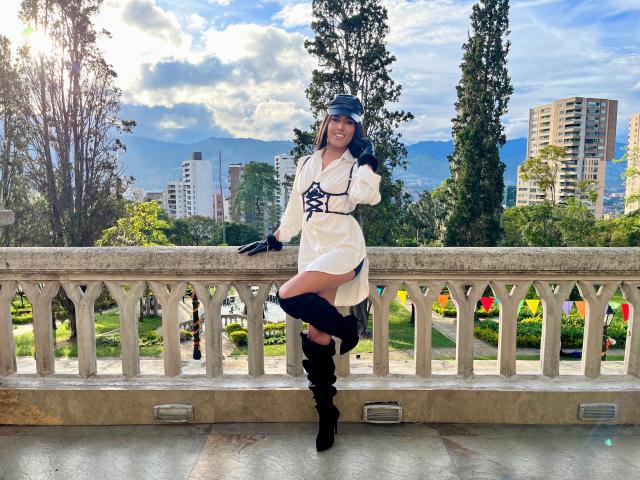 FallonRogers, avec des cheveux bleus, une robe blanche, des bottes noires et des gants, appuyée sur un balcon en pierre, souriante, avec des montagnes et une ville en arrière-plan, baignée de soleil, ciel clair..