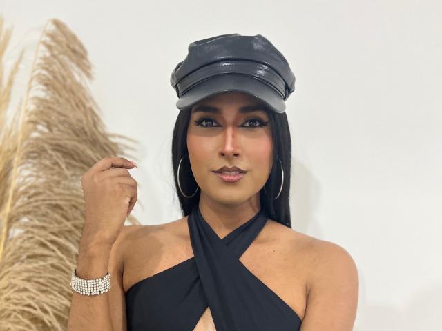 FallonRogers con una gorra de cuero negro, aros grandes y una blusa negra con cruces, mostrando sus pequeños pechos y su linda sonrisa, llevando una pulsera de diamantes, con hierba alta de color beige..