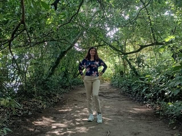 BulmaHot staat op een zandpad, gekleed in een blauw bloesjemmotief shirt, beige broek en witte sneakers, met haar handen in haar zakken, omringd door groene begroeiing en hangende takken..