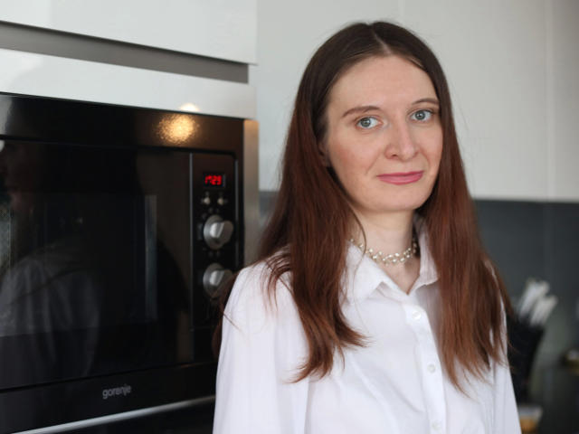 DannyDevil-hot has straight brown hair, blue eyes, and is wearing a white button-up shirt with a pearl necklace. She stands in front of a black microwave.