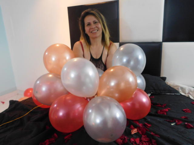StarBebes mit blonden Haaren, lächelnd, in einem schwarzen Spitzenoberteil, sitzen auf einem schwarzen Bett und halten rosa, silberne und rote Luftballons, Blütenblätter sind auf dem Bett verstreut..