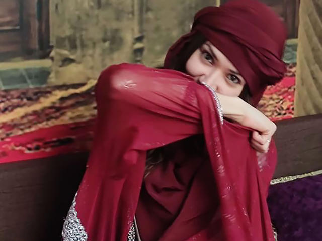 SujannaVarma with red headscarf, hugging a red sheer garment over her tits, wearing a patterned top, looking shy. Her brown eyes peek out. Middle shot.