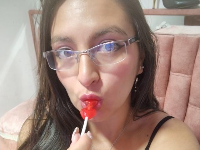 VenusSweetPalmer with glasses, sucking on a red lollipop, holding it with her fingers, pink lips slightly parted, dark brown hair, wearing a black spaghetti strap top.