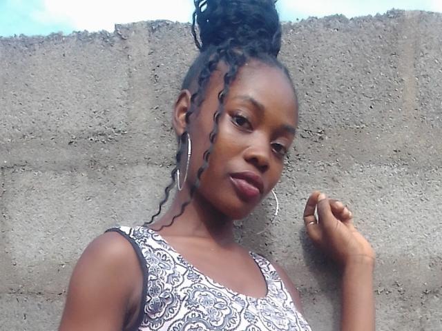 EveHot with dark skin, curly black hair in a high bun, wearing a patterned sleeveless top, hoop earrings, and a ring, standing against a concrete wall.