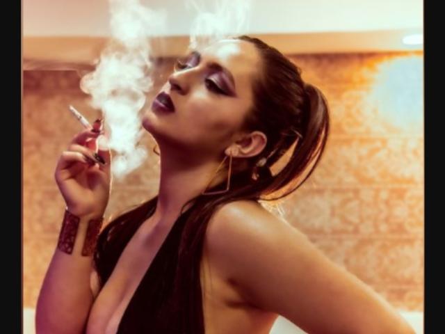 VenusSweetPalmer con maquillaje ahumado, labial oscuro, top de tirantes negros, sosteniendo un cigarrillo, humo elevándose, coleta alta, aretes dorados, pulsera roja, sexy, seductora, piel bronceada, a medio desnudo..
