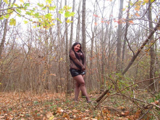 EmmaHotSquirt-sex with long dark hair, wearing a black sheer top and black leather shorts, standing in a forest with leaf-covered ground and bare trees.