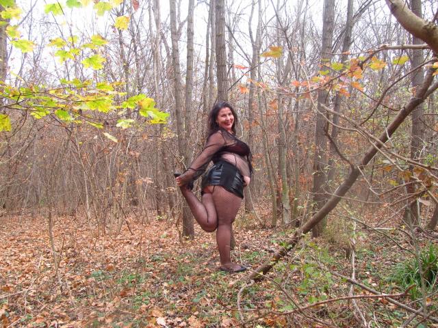 EmmaHotSquirt-sex stands in a forest, lifting one leg, wearing black sheer stockings, a brown top, and black leather shorts, with a playful smile and curly dark hair.