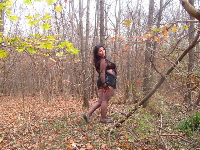 EmmaHotSquirt-sex standing in a forest, wearing a brown top, black shorts, and purple tights, looking back with a slight smile, leaves on trees, and a fallen branch.