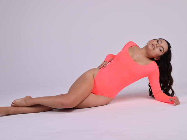 JasmineAsha is lying down in a neon pink leotard, legs stretched out, back arched, hand on hip, and head tilted to the side, looking relaxed and sexy.