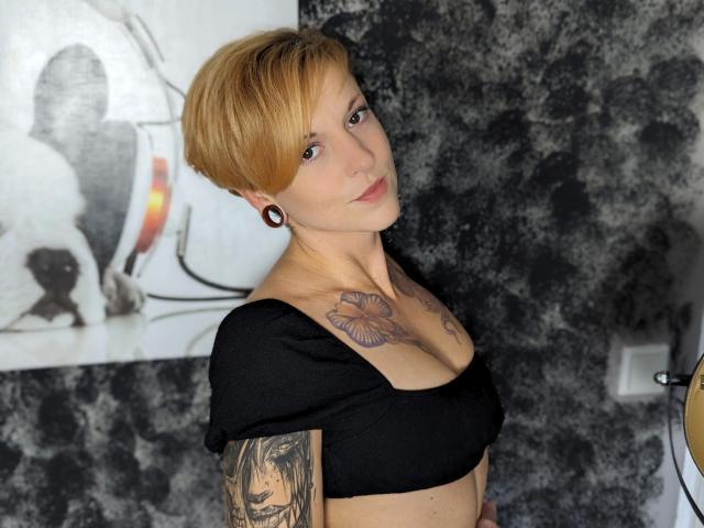 MiaSonne-hot with short blonde hair, black crop top, tattoos on chest and arm, black earbuds, looking at camera, gray textured wall, white wall switch on right.
