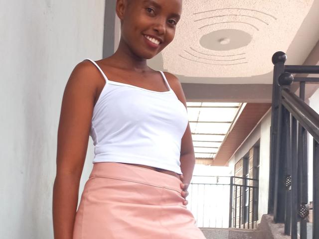 Africanah with dark brown skin, small tits in a white tank top, and a pink skirt, smiling, standing on a balcony with black railings, hand on hip.