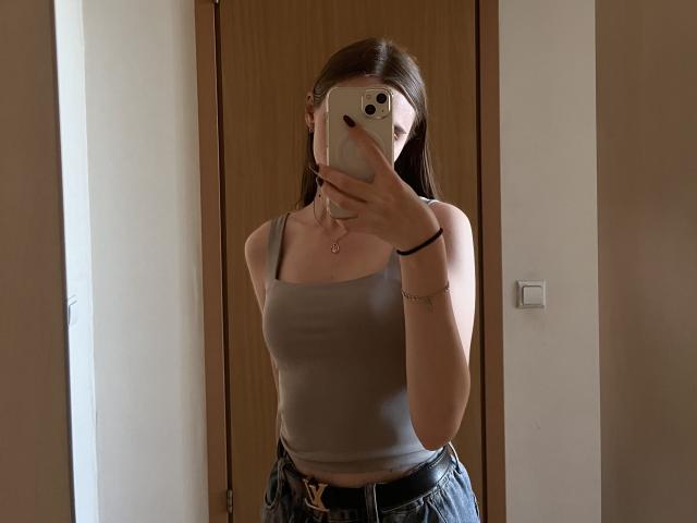 Valissia with medium tits, wearing a grey tank top and black jeans, taking a mirror selfie with a phone, brown hair, black bracelet, and a pink necklace.