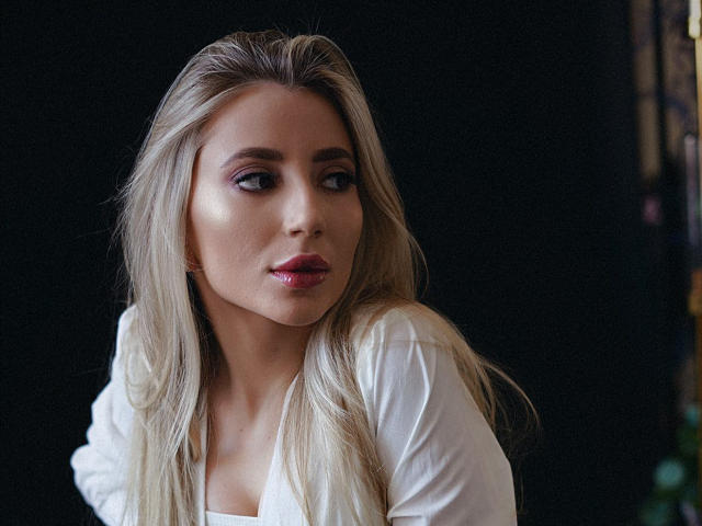 NicoleNovak with long blonde hair, white shirt, subtle pink eyeshadow, glossy pink lipstick, looking slightly to the side, dark background, soft lighting.