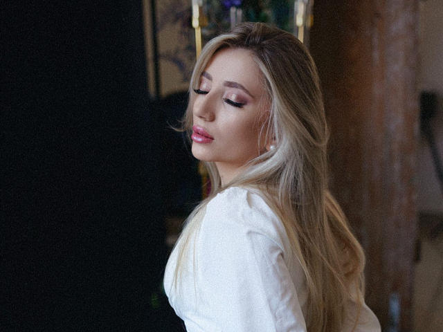 NicoleNovak has blonde hair, pink lipstick, and closed eyes, wearing a white shirt, with soft light highlighting her smooth skin and slightly parted lips.