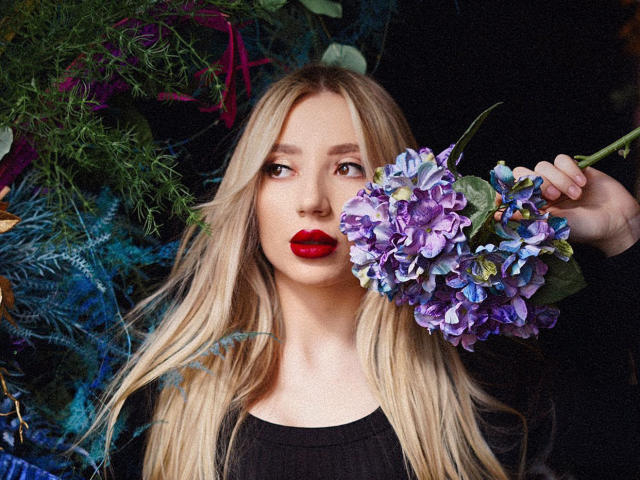 NicoleNovak with long blonde hair, red lipstick, holding a purple and blue hydrangea, wearing a black top, looking to the side, in a dark, leafy background.