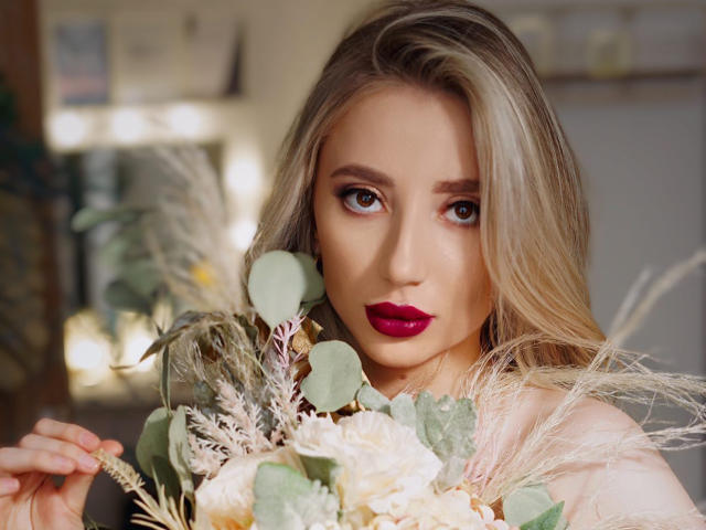 NicoleNovak with blonde hair, red lipstick, holding a bouquet of flowers, green leaves, and white pampas grass, looking seductively at the camera.