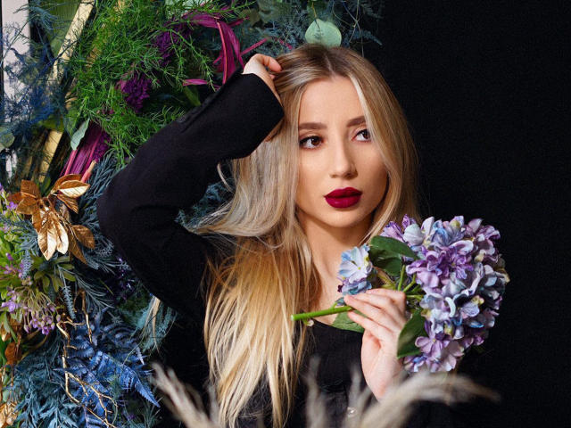 NicoleNovak with long blonde hair, red lipstick, black top, holding purple hydrangeas, hand in hair, surrounded by colorful foliage and flowers, looking to the side.