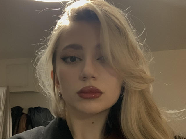 NicoleNovak has blonde hair, a cute face, and shiny red lips. Her left eye is winking, and she's wearing a black top with a collar. Her ass is not visible.